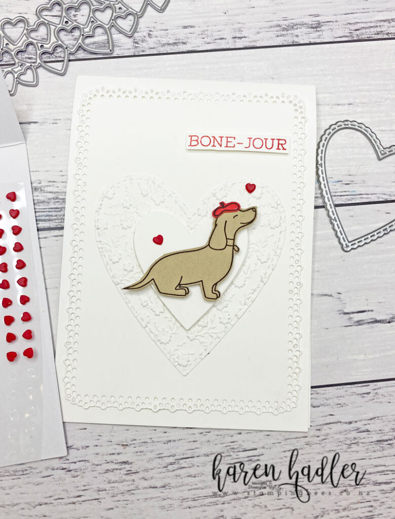 A White card with a White Mat and two white hearts. There is a Brown Hot Dog in the middle with a red Beret on . The are words stamped in red saying Bone Jour top righ hand third of the card. There is also 2 little red hearts.