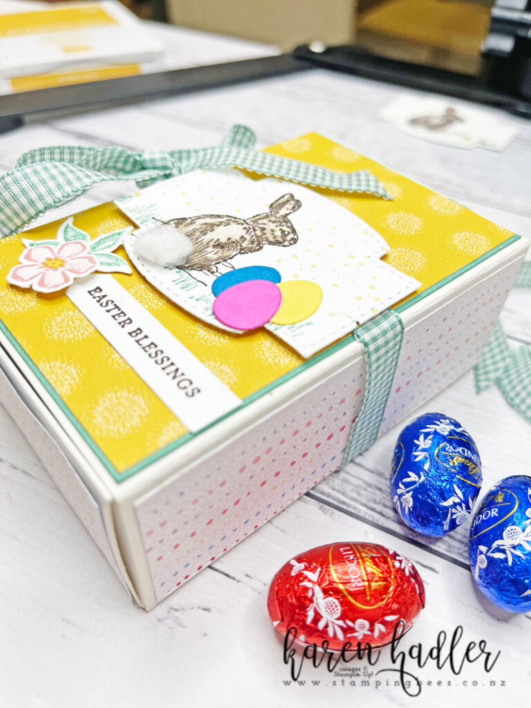 A treat box full of mini Easter Eggs Decorated with a green Gingham Ribbon. On the front is some yellow DSP a cute brown Bunny with a fluffy white tail. Next to it is 3 little bright paper easter eggs one in Pink, blue and yellow. The sentiment at the bottom says Easter Blessings and has a little flower with a leaf to the left of it.