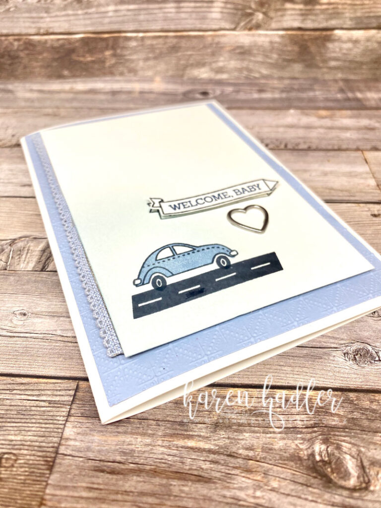 A close up ofA welcome bay Card on a White base with an embossed blue mat. On top of this is a white mat and a blue scalloped ribbon running down the left hand side. In the bottom left hand corner on top of some soft blue blending is a blue car driving on a black rd. There is a banner which says Welcome baby and a little silver heart below.