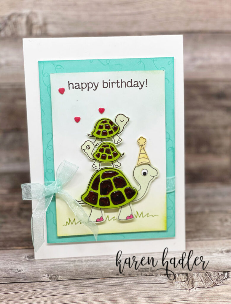 Turtle friends. a Happy Birthday Card set on a Coastal Cabana Mat. The background is sponged blue for the sky and green for the grass. There is a large green Turtle on the bottom with two smaller turtles stacked on top. The large turtle has pink toenails. The is a sheer pool party bow on the left.