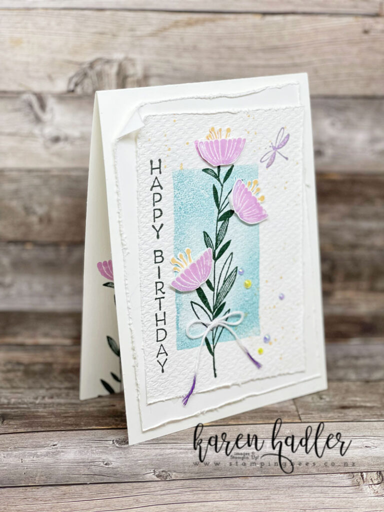 Celebrate with Flowers with Water Colour Shapes Stamp sets. The background is stamped in Balmy blue with a vertical rectangle. The flower stem is stamped in green running up the middle. The Happy Birthday is also vertical and co-ordinates in green. The flowers are in Fresh Freesia with 3 cut out and on dimensionals. There is a white card base and mats given texture by the edges being roughed up and the top stamped layer being run the with the tasteful texture embossing folder.