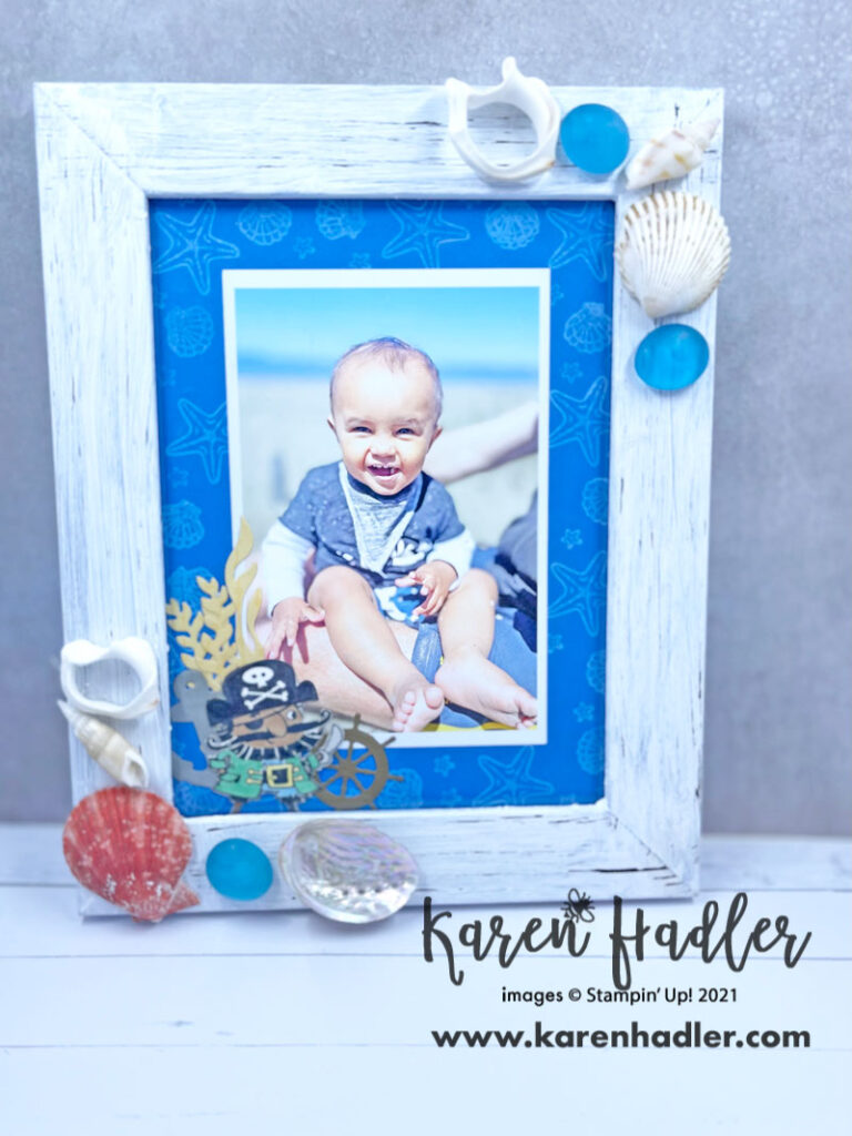 A white distressed picture frame with Shells top right and bottom left corner. the matting inside the frame is a vibrant blue with white shells stamped around the outside. In the bottom left hand corner i a cute pirate with an anchor , Seaweed, and a steering wheel.