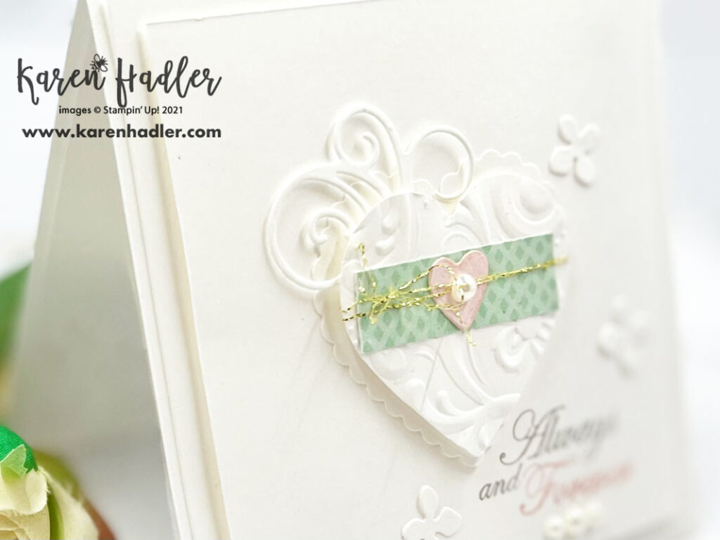 A close up ofForever Blossoms a White square card with a swirled embossed heart on the front. There is die cut swirls coming out of the top left of the heart and small white flowers scattered across the front. In the centre of the card is a small strip of mint Macaroon DSP and a small pink heart. Wrapped around these and the heart is some gold thread tied in a bow. The Sentiment reads Always and Forever.