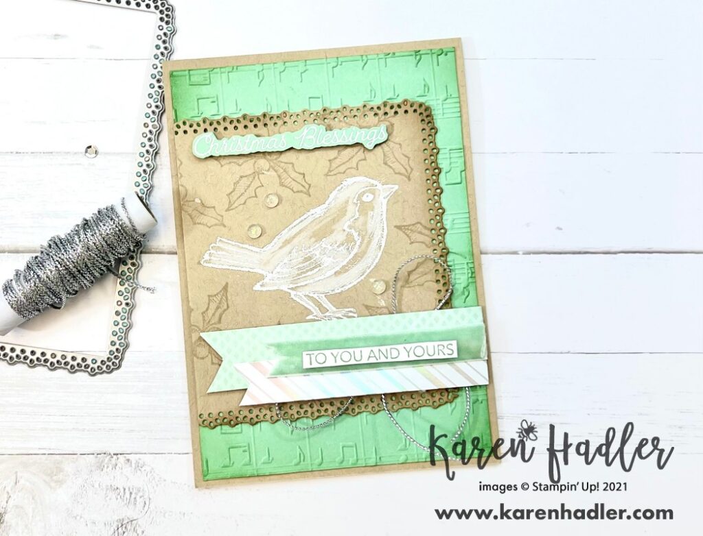 Happy Holly-Day Card with Crumb Cake base and Mint macaroon matting. The mint macaroon mat has been embossed with music notes. On to with a Lacey edge is a crumb cake mat and this has the bird white embossed on the front. This bird has then been coloured in only white. The background has soft Holly Leaves. At the top white embossed on Mint macaroon is Christmas Blessings. There are strips of Mint macaroon DSP and ribbon going right to left along the bottom and the sentiment on top is to you and yours. Stamped with Mint Macaroon on White card Stock.