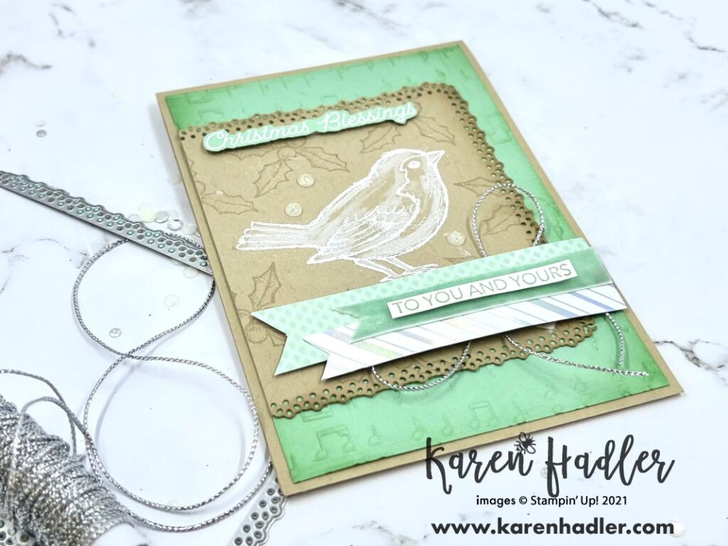 A closer look at the layers of Happy Holly-Day Card with Crumb Cake base and Mint macaroon matting. The mint macaroon mat has been embossed with music notes. On to with a Lacey edge is a crumb cake mat and this has the bird white embossed on the front. This bird has then been coloured in only white. The background has soft Holly Leaves. At the top white embossed on Mint macaroon is Christmas Blessings. There are strips of Mint macaroon DSP and ribbon going right to left along the bottom and the sentiment on top is to you and yours. Stamped with Mint Macaroon on White card Stock.
