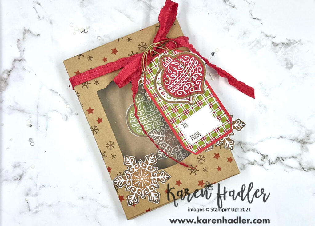 A picture of the Kraft Gift box stamped with red stars and brown snow flakes. There is a red ruffled ribbon tied around the top in a bow. From that is a tag using the Gingerbread & Peppermint DSP as the base and elements such as baubles and large snow flakes have been fussy cut to decorate.