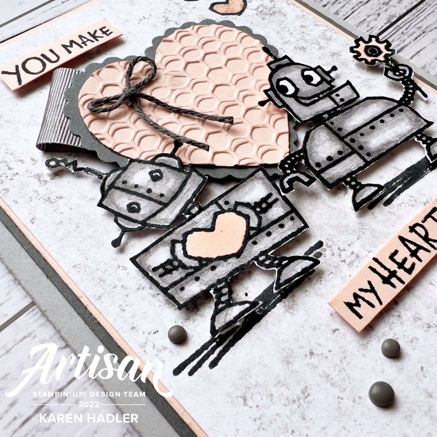 A close up ofA picture of a greeting card with text. The colours are pink and Gray. There is a Large  pink heart in the middle of the card with two cute Robots either side. One has a heart on his chest and the other is holding a flower. The text reads You make my heart go Beep
