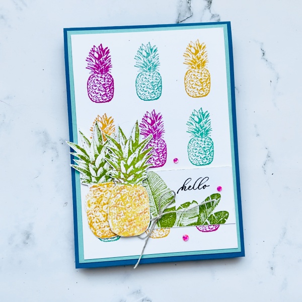 A picture of a card with Text. There is many small pineapples in the background. Bright Pink, Blue and yellow. The are two large bright yellow pineapples and the text reads Hello