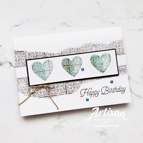 A picture of a greeting card with Text that says Happy Birthday. The colours are white, brown and blue. There is a tile pattern underneath 3 blue pattered hearts in the middle. with a small bow.