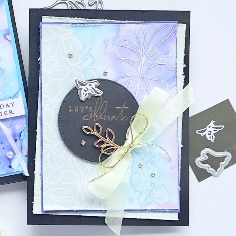 A picture of a card showing the Water colour Emboss Resist Technique in a soft background  with purples, greens and pinks. The sentiment reads Let's Celebrate