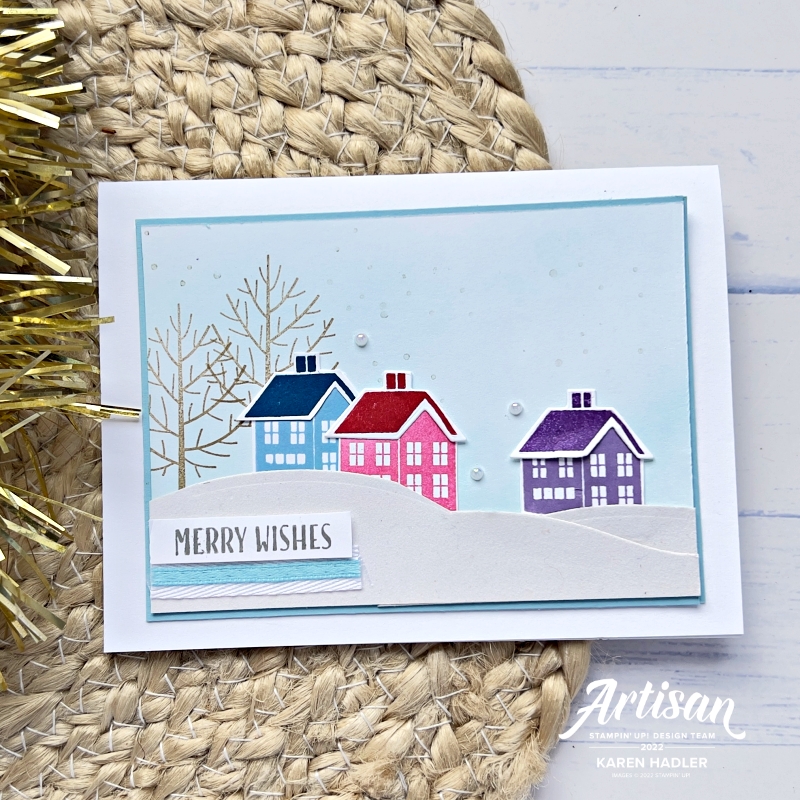 A picture of a Christmas Greeting Card with a Blue sky with Snow Falling. 3 brightly coloured houses sitting on top of a snowy Hill. The sentiment reads Merry Wishes