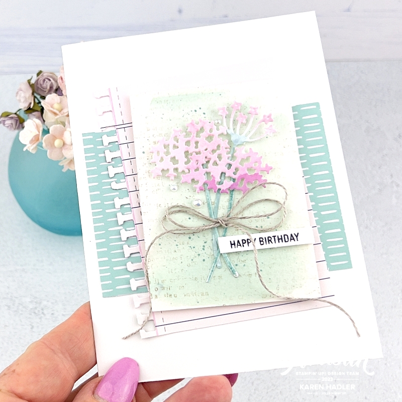 A picture of a greeting card in the centre is a water coloured piece of card in Pool Part on top of this is  pink water coloured flowers that look a little like hydrangeas. the sentiment reads Happy Birthday and there is a large double linen thread bow. The background has a piece of co-ordinating Pool party paper with small white lines. On top of this is a pice of paper that looks like it is torn from a notebook.