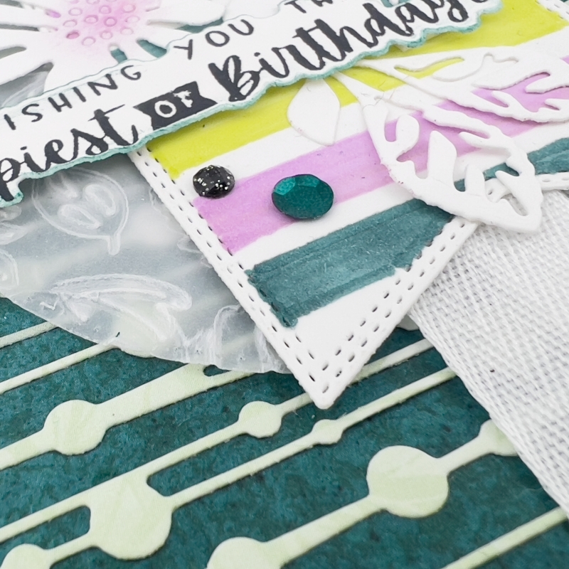 A close upA picture of a Birthday Card. In the background the is stripes and spots. In the foreground there is a tag with, pink, green and yellow stripes. On top of this there is 2 daisies with pink centres. The sentiment reads wishing you the Happiest of Birthdays