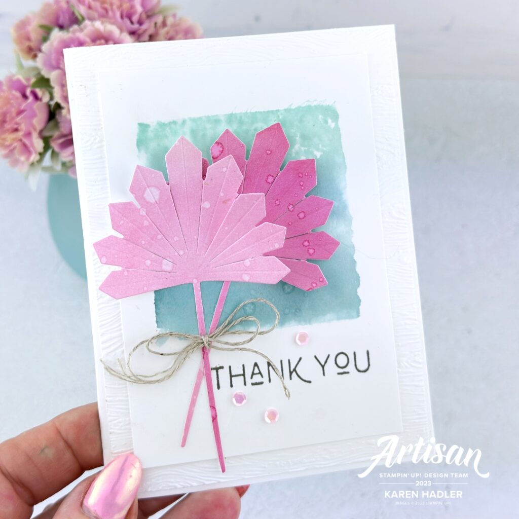 A picture of a grfeeting card that says Thank You. It is a Basic White Card Base with a water coloured green Blue square in the middle. On top of this is 2 Pink palm leaves that are tied with a linen bow.