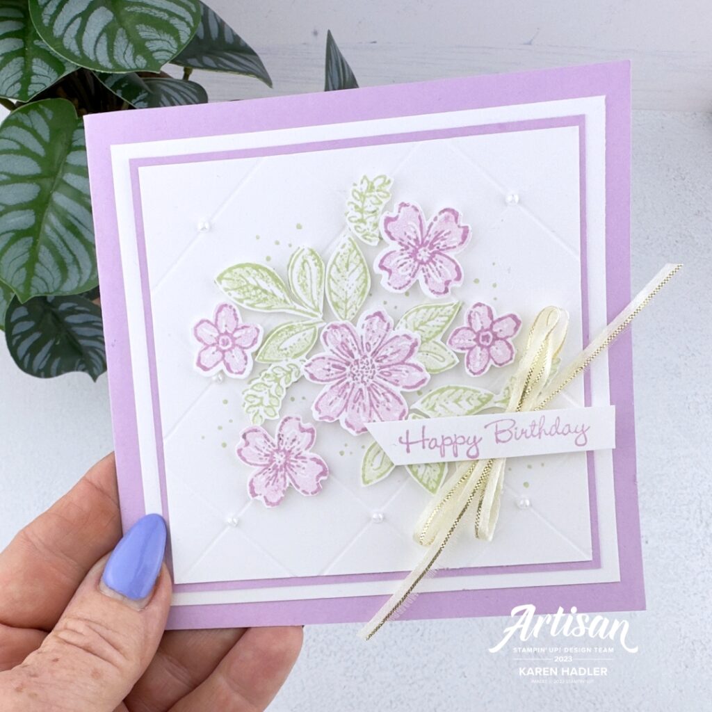 A square fresh Freesia Greeting card that reads happy Birthday. On the front is a white panel with a criss crossed embroidery parttern. On top of this is some Fresh Freesia Flowers and Green leaves. There is a large gold bow under the sentiment and pearls at every cross on the background.