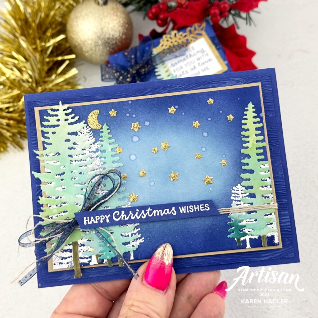 A Starry Sky Blue Christams card that depicts a night scene with gold glittery stars and moon. To the left and right are white embossed trees and on top of the these are some green water coloured tress. The sentiment is white embossed of match Starry Sky blue card stock and reads Happy Christmas Wishes. The is a blue glittery bow to the left.