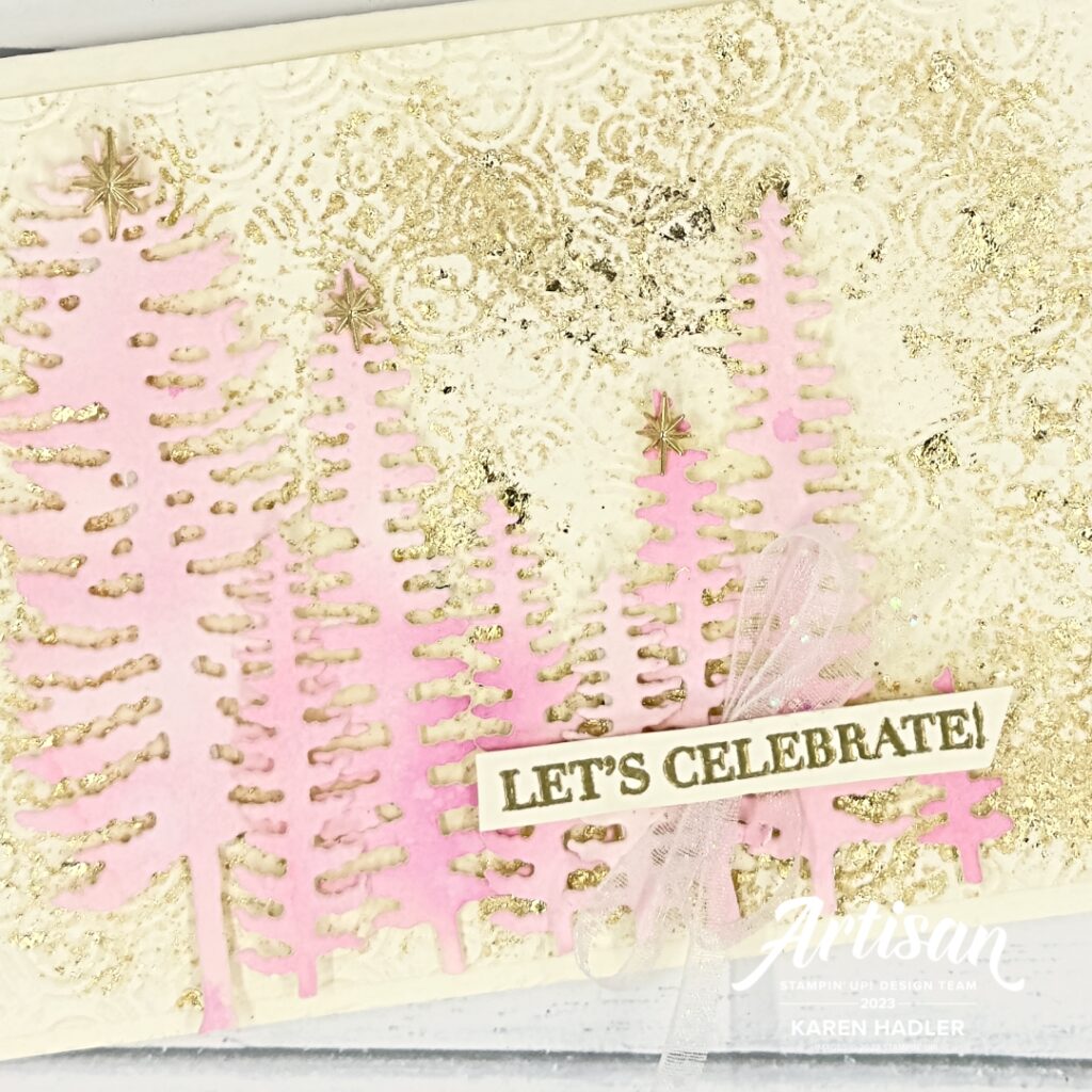 A close up ofA greeting card that reads let celebrate. The background is embossed with a distressed Tile embossing folder and there is gilded leafing randomly placed. on top is a row of pink water coloured trees . The sentiment is gold embossed across the top of the trees and underneath is a small iridescent bow.