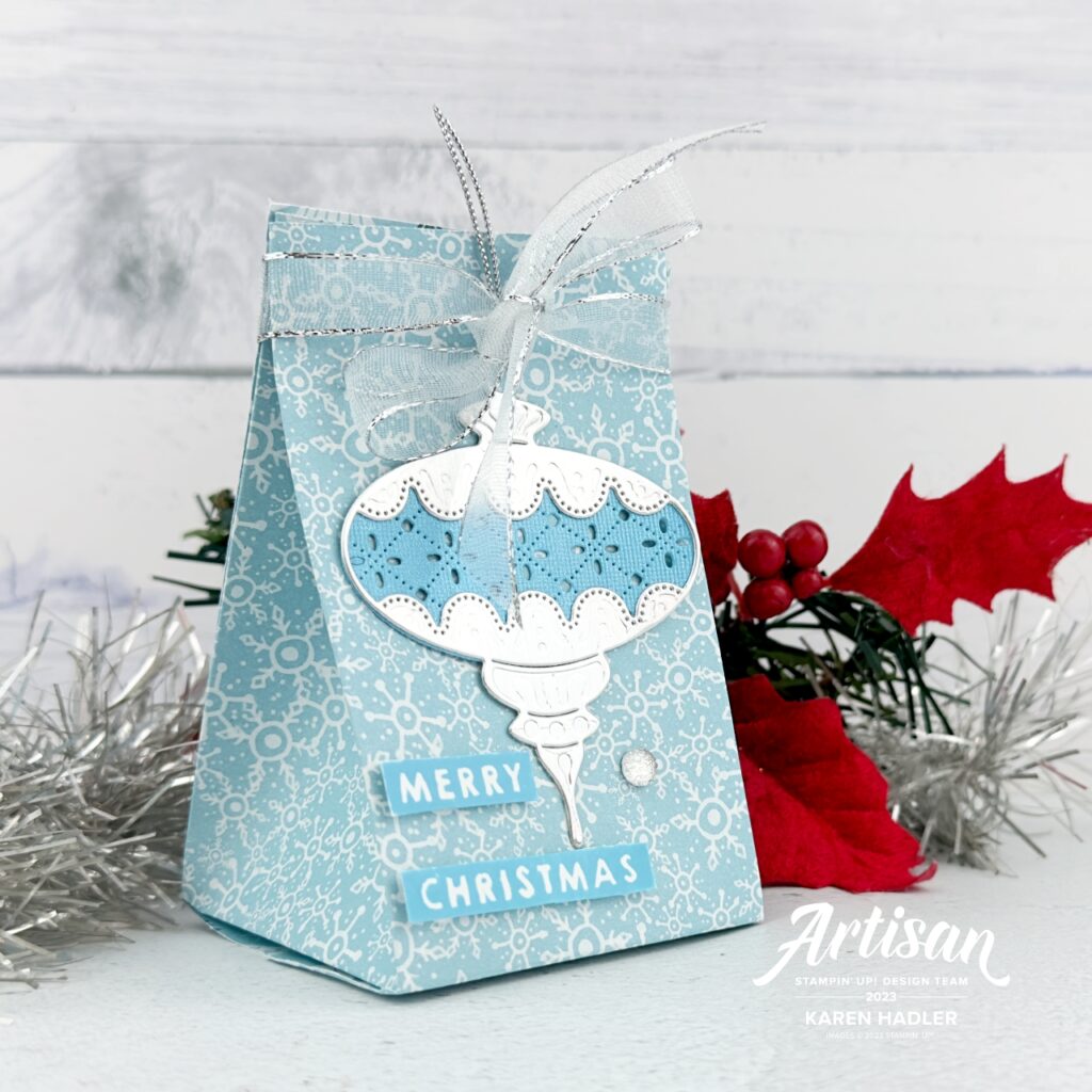 A small handmade gift bag made of blue paper with small green and white trees.  on the front is an Ornament decoration that is Silver and Blue There is a silver and White bow at the top. The sentiment reads Merry Christmas. 