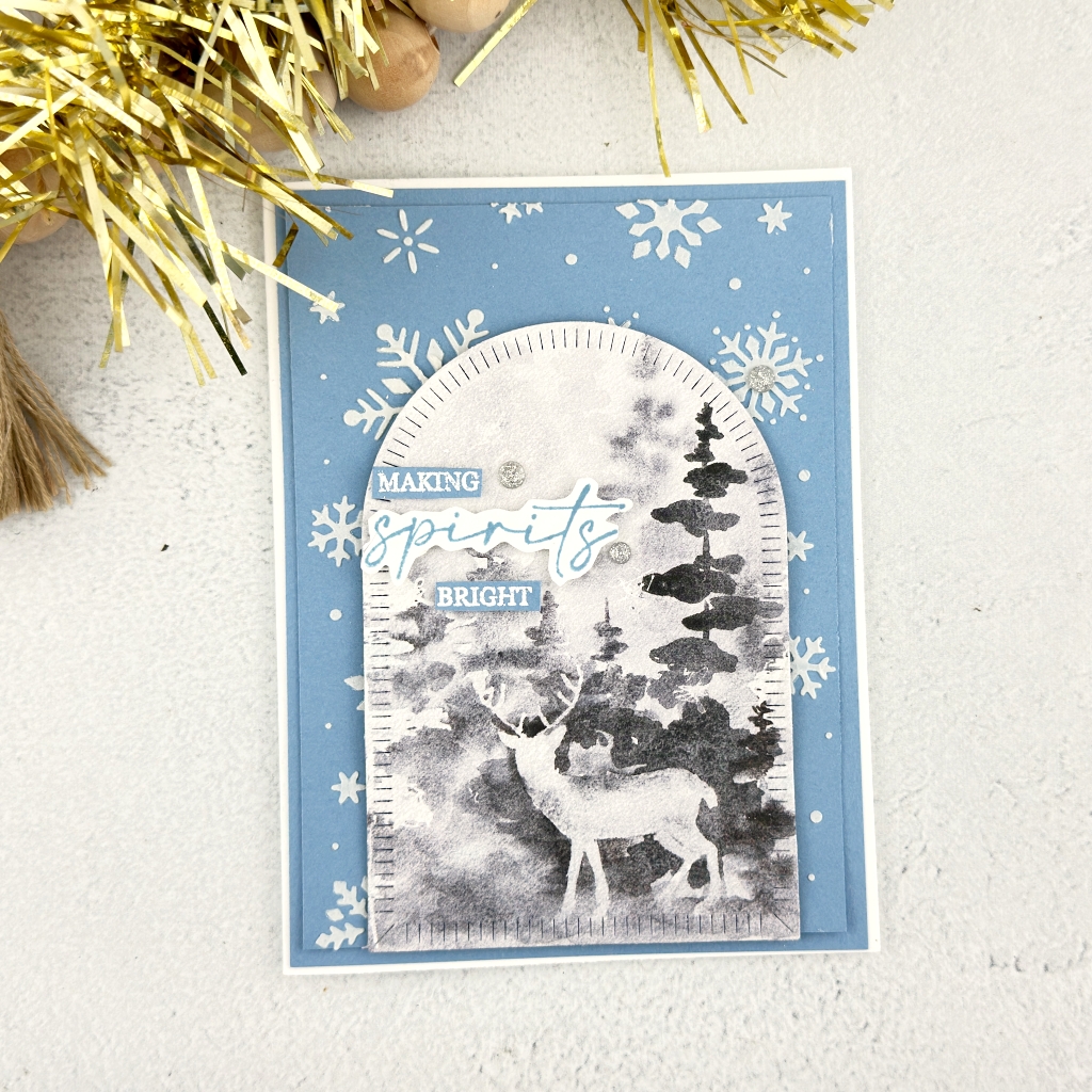 A Blue Card with White Shimmery Snowflakes in the background, in a doorway sahpe is an image of a deer in aforest at dusk. The sentiment reads Making Spirits Bright 