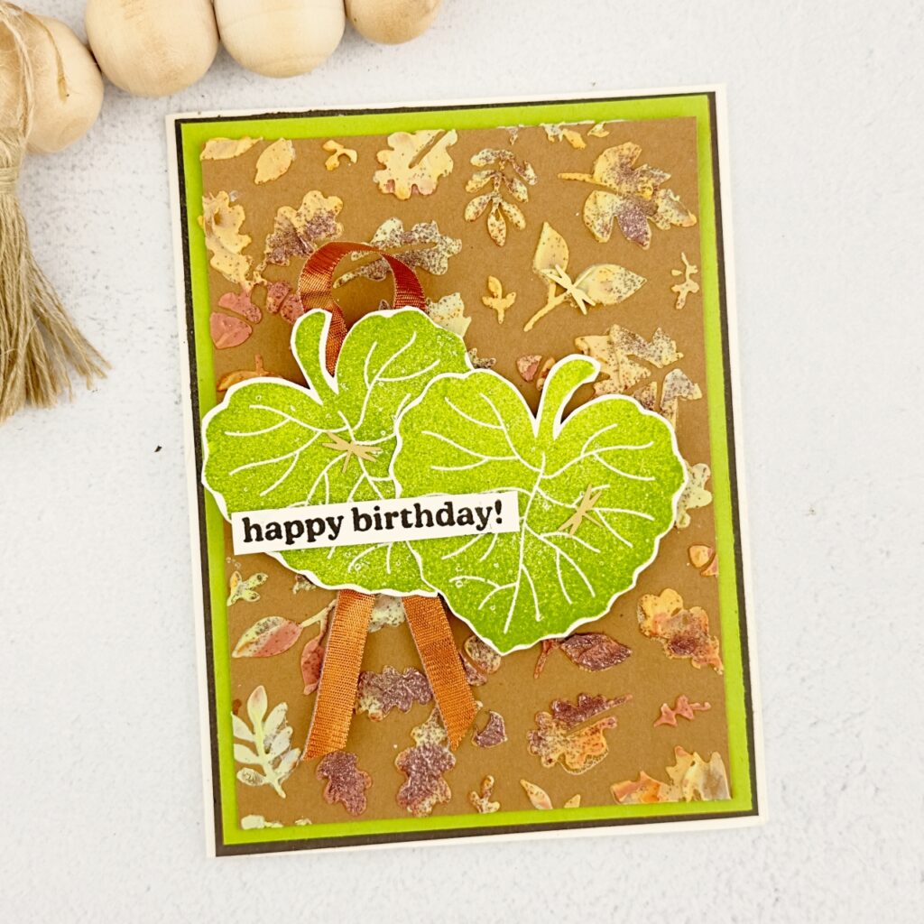 A greeting Card on a green back ground the front is a light brown with Autumn coloured leaves. The are two large green leaves with a copper ribbon underneath. The sentiment reads happy birthday. 