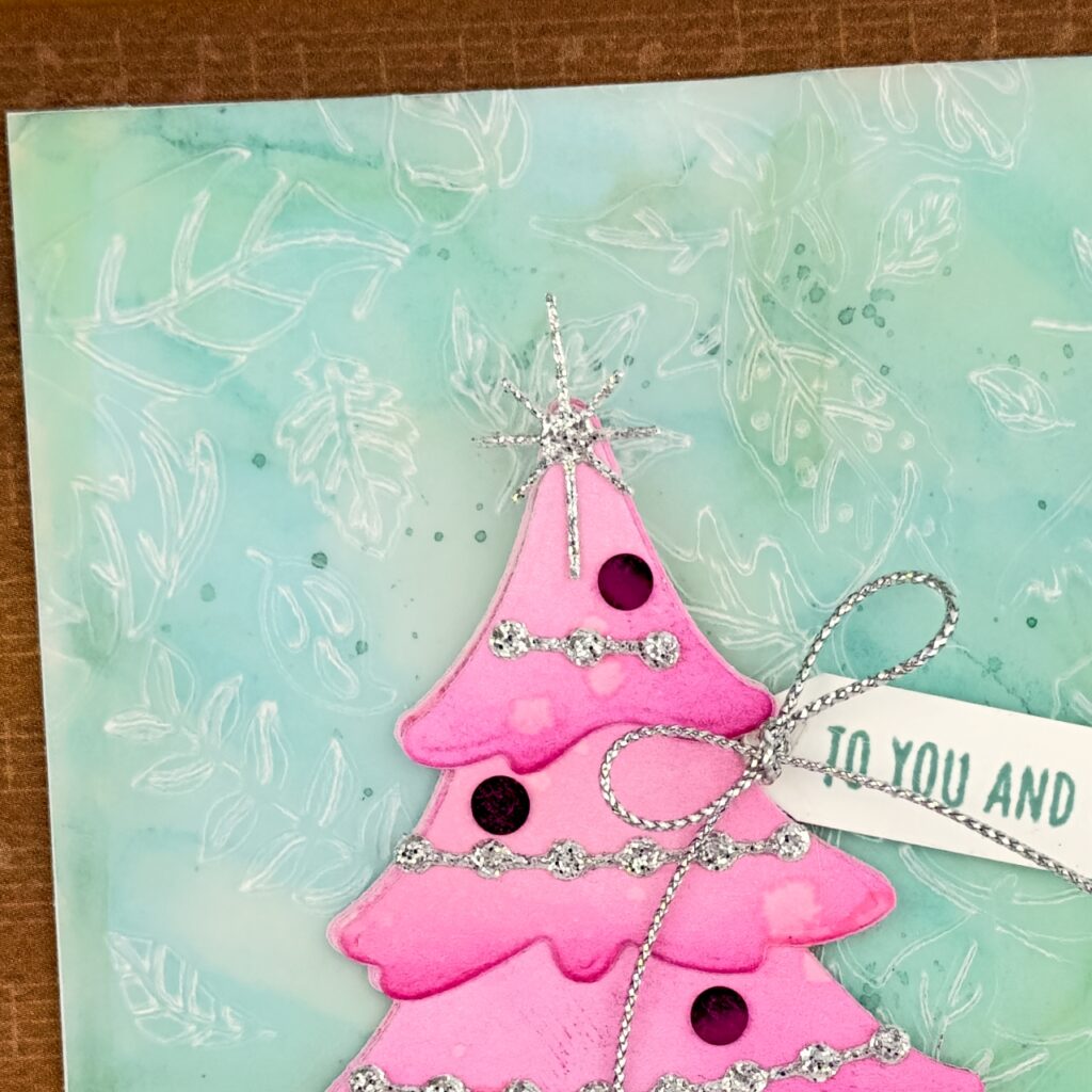A close up ofA greeting card on a Pecan Pie card Base. Ontop of this is a mat in a similar colour with musical notes on it. On top of this and smaller is a white card mat with a green coloured and embossed piece of vellum. The patter is leaves. On top of this is a large pink layered Christmas tree with silver and pink decorations. The sentiment is to You and Yours in the form of a gift tag and has a Silver Trim bow. 
