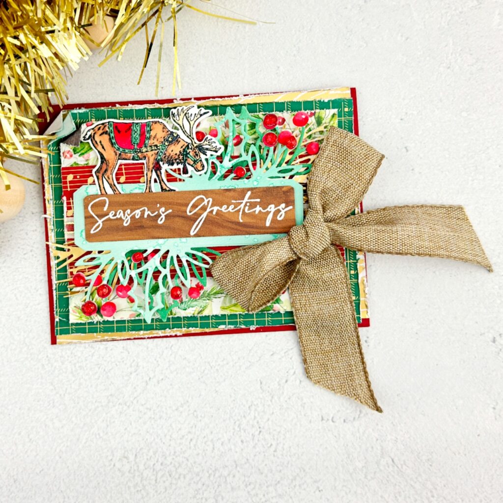 A Christmas card that has many layers of designer Series paper. Gold, green and red each piece has been roughed up around the edges. On top of this is a label that is two shades of green the darker one being on the bottom. This label as holly leaves and berries all around it. On top of the label is a wooden piece of DSP that has white embossed on it Seasons Greetings. On top of the label is a Regal Christmas Reindeer . To the right of the label is a large luxurious bow tied in a natural coloured material.