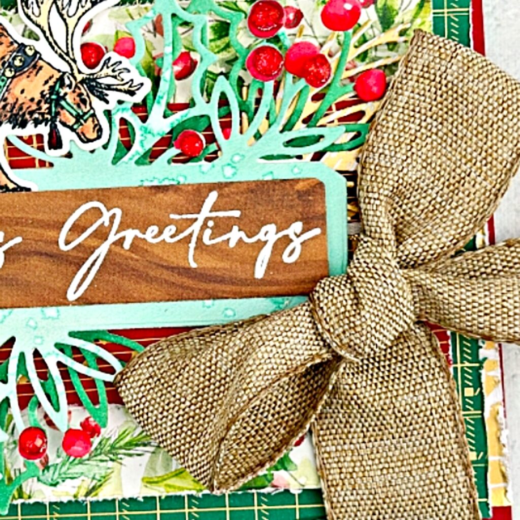 A close up of A Christmas card that has many layers of designer Series paper. Gold, green and red each piece has been roughed up around the edges. On top of this is a label that is two shades of green the darker one being on the bottom. This label as holly leaves and berries all around it. On top of the label is a wooden piece of DSP that has white embossed on it Seasons Greetings. On top of the label is a Regal Christmas Reindeer . To the right of the label is a large luxurious bow tied in a natural coloured material.