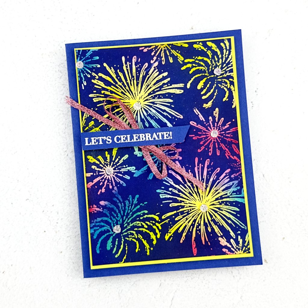 A greeting card that has a dark blue Starry Ky base a mat of lemon Lime Twist. On top is a multitude of bright coloured fireworks on a blue background in blues yellows and pinks.The sentiment which is going from the left hand side of the card is white embossed on the blue cardstock and reads Let';s celebrate. Underneath\h is pink woven ribbon tied in a bow. There are silvery embellishments in the centre of each firework
