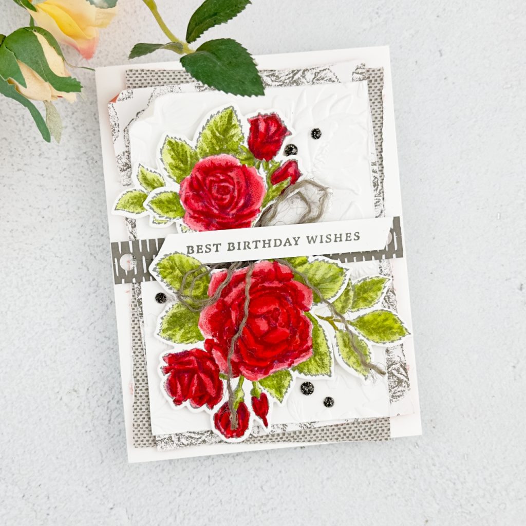 A Birthday card on a Basic White card base on top of this is 2 mats with different patterns on it in Pebbled path adhered at different angles. Ontop of this is an embossed piece of white card stock. The embossing is flowers. Across this is a strip of Pebbled Path Paper with small white stripes and small circles. There are to large red roses some red rose buds and green leaves going from top to bottom. Across the middle is a large bow in jute thread in pebbled path and the sentiment on top of this which reads Best Birthday Wishes