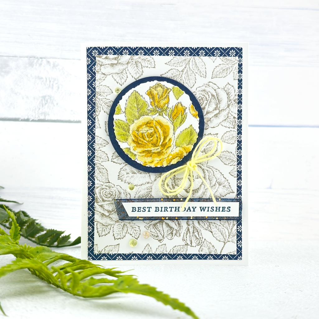 A card on a white card base. the next mat is a navy and blue patterned piece of DSP on top of this is a piece of White card stock stamped with roses and leaves. A circle as been cut out of this pattern near the top coloured placed on a blue mat placed on dimensionals and put back in the same place. This is called the spotlight technique. There is a yellow below the circle to the right. Underneath this is a strip of white card with Best Birthday wishes stamped in blue.. 