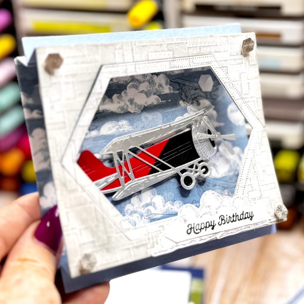 A greeting card that is a mini Shadow Box the base is boho Blue and the front has gray riveted patterned paper. In the for corners are embellishments that look like studs. The middle is cut out and looking inside you can see cloudy skies with a large old fashioned biplane flying across the sky. The plane is red and black with silver wings.
the sentiment is on the front ontop of a small purple cloud and reads happy Birthday