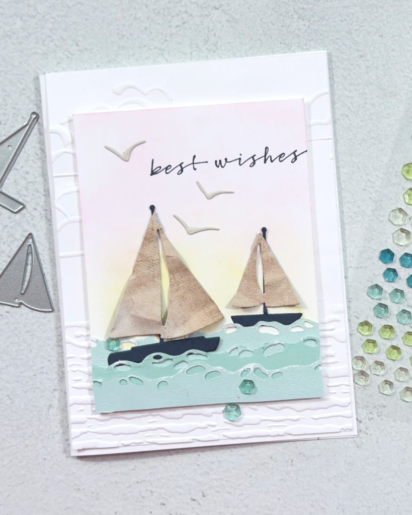 A white based greeting card with a smaller square on the front that has been blended with yellow and pink for a sunrise. In the for ground there are 2 sailing boats in the waves. There a re seagulls and the sentiment is stamped on the card from and reads Best wishes.