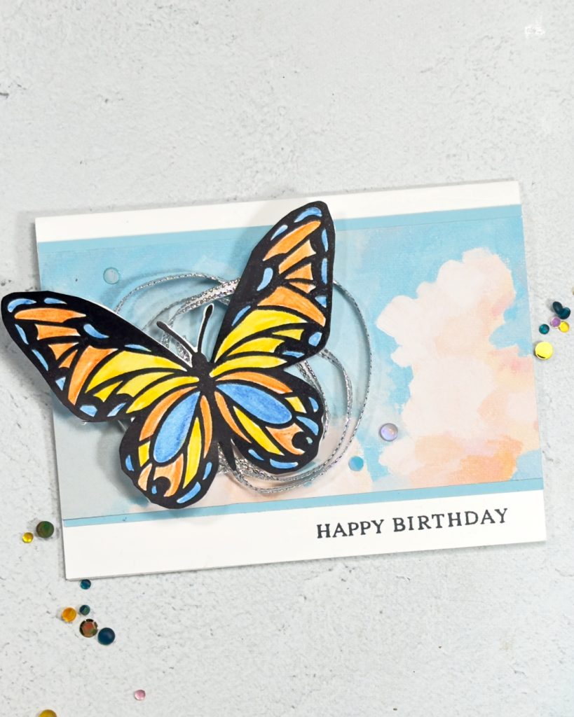 A greeting card on a white base. The background is a cloudy sky and on the left is a large brightly coloured Butterfly. The colours are blue, yellow and orange. Underneath the butterfly is silver twine that has been wrapped around in a couple of circles.
The sentiment reads happy Birthday at the bottom right.