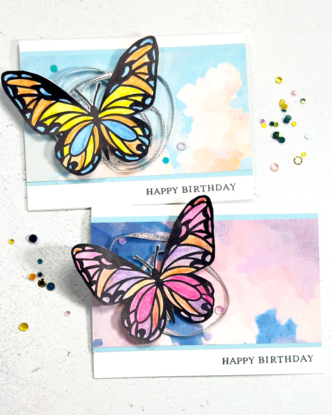 Two greeting cards on a white base. The background is a cloudy sky and on the left of each card is a large brightly coloured Butterfly. Underneath the butterfly is silver twine that has been wrapped around in a couple of circles.
The sentiment reads happy Birthday at the bottom right.