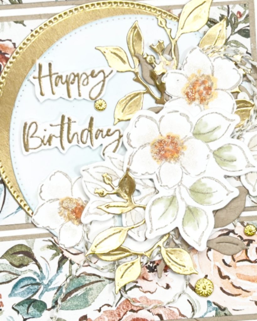 A close up ofA floral Birthday card on a white base. With Gold and sparkle. There is a gold and white circle in the middle with a posy of flowers on the right hand side . The sentiment is gold embossed inside this circle and which also has gold leaves.