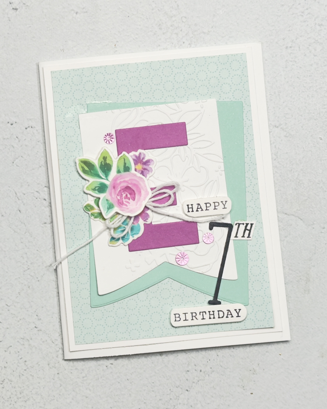 A childs age 7 Birthday card for a girl in Pinks and Pool Party There are 2 banners off set on the front with a large letter E with flowers adorning the middle. The sentiment reads Happy Birthday.