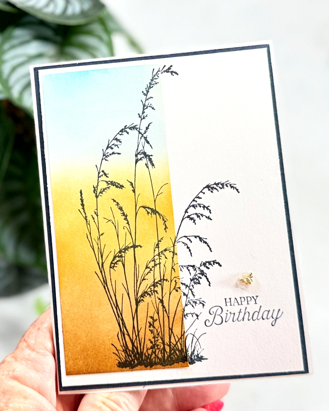 A white based Birthday Greeting card that has a brown gold and blue strip of colour down the left handside and on top in black some stamped grasses. There is a tiny gold bee above the sentiment which is bottom right.