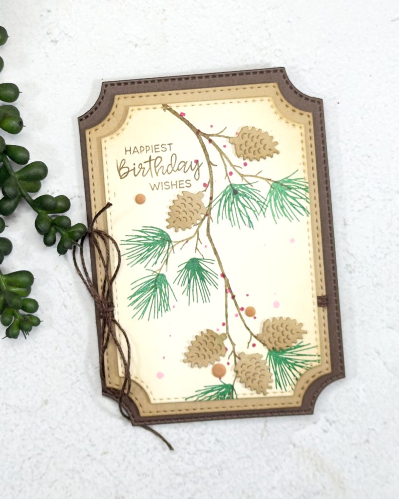 An early espresso and crumb cake layered birthday card.There is a long branch coming down from the top with pine cones and greenery that looks like pine needles.