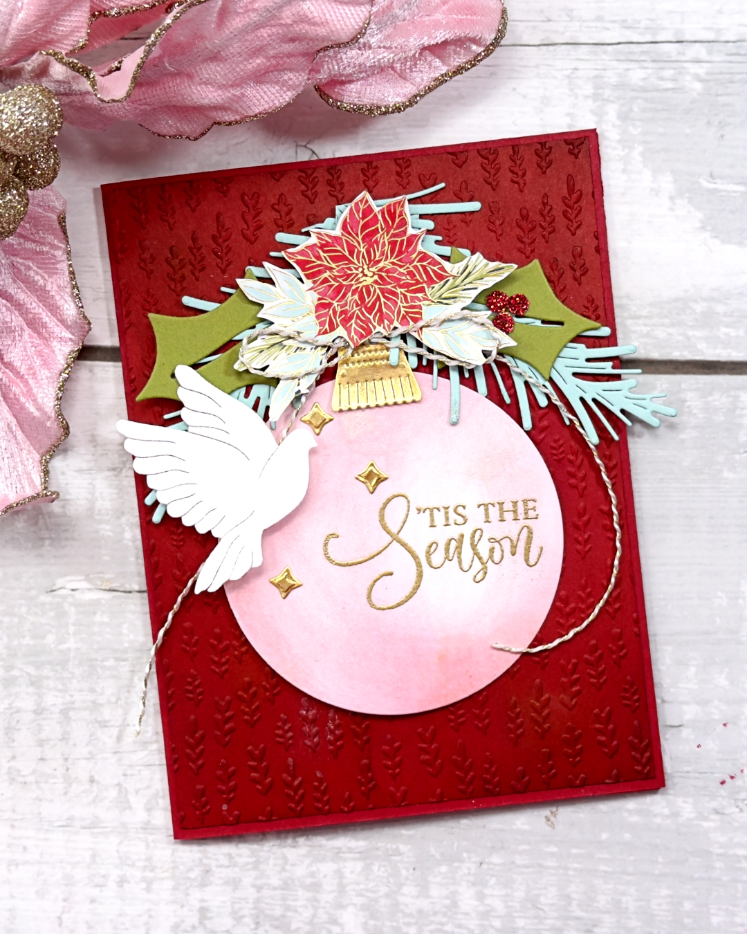 A red Christmas greeting card with a large pink bauble as the feature, The sentiment 'Tis the season is gold embossed on the bauble. There is a poinsettia nd greenery on top pf the bauble and a white dove to the left.