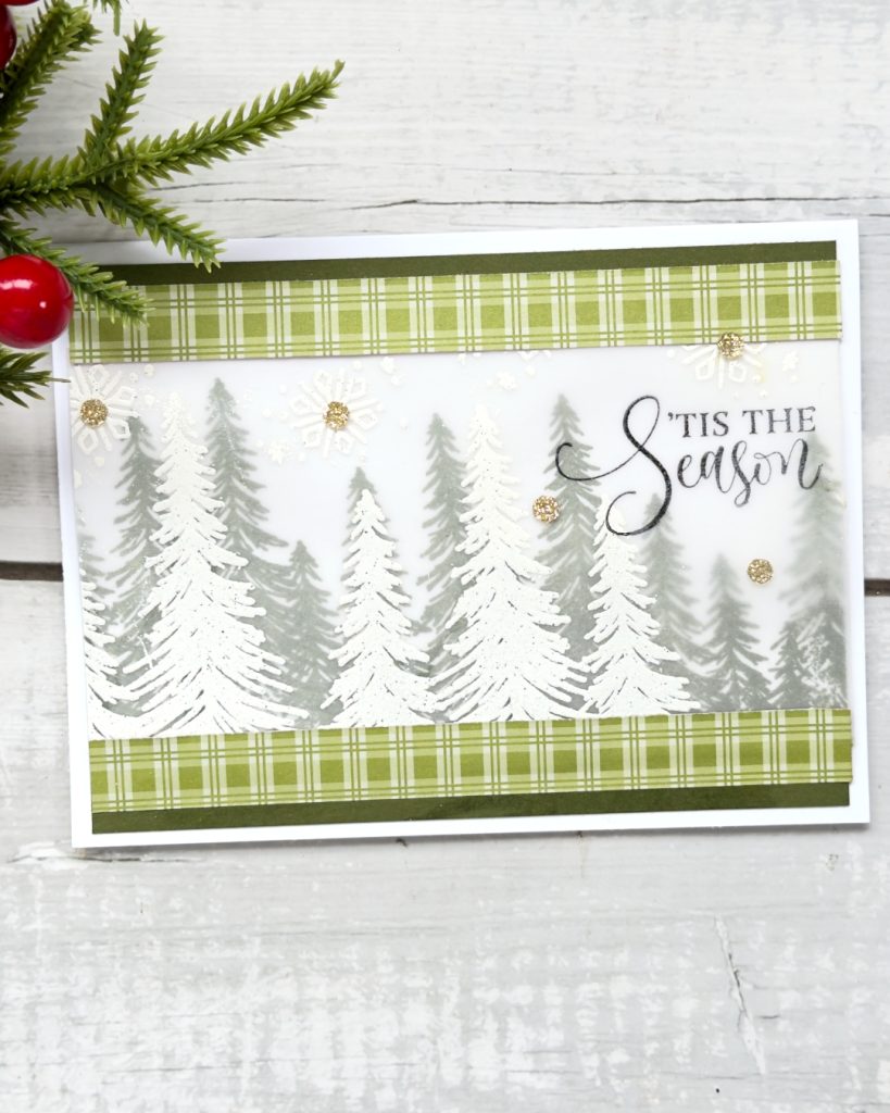 A green coloured horizontal Christmas card with trees through the middle. A strip of plaid aper top and bottom. the trees are covered with vellum which has white embossed trees on the top.and the sentiment reads 'Tis the season