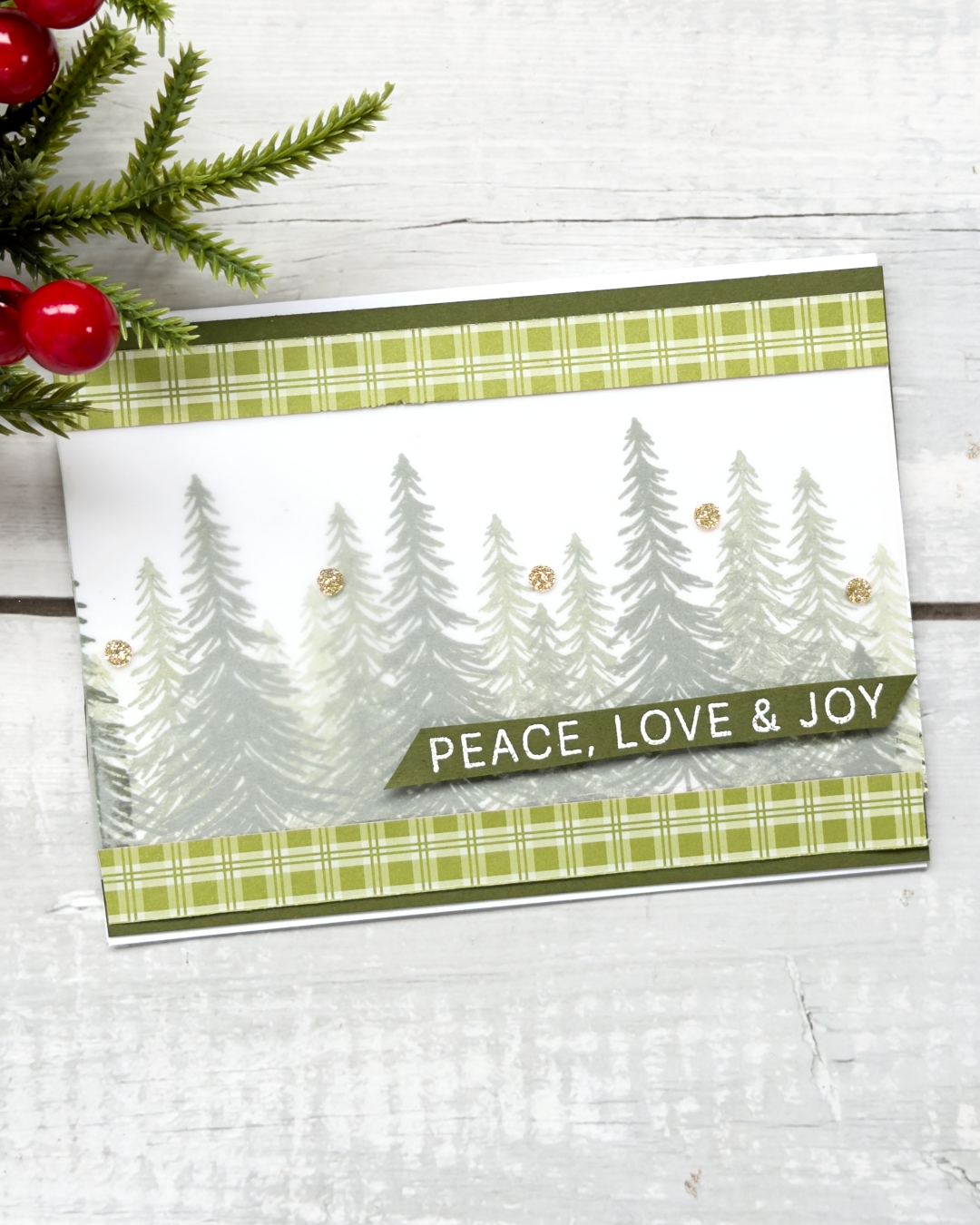 A green coloured horizontal Christmas card with trees through the middle. A strip of plaid aper top and bottom. the trees are covered with vellum and the sentiment reads Peace,love & Joy
