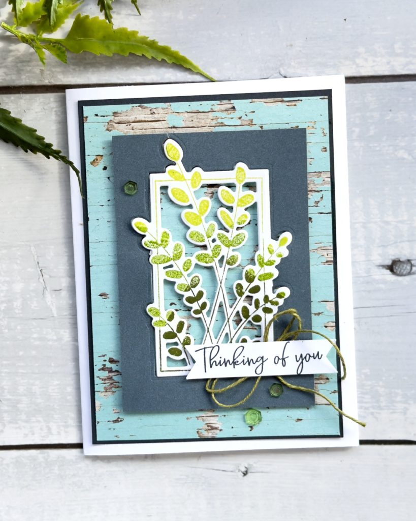 A greeting card thats sentiment reads Thinking of you. It is on a White Card base with some blue wood pattern matting. In the centre is a posy oif green leaves there is a double bow and embellishments.