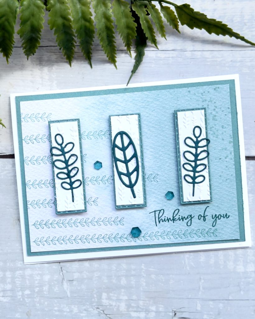 A greeting card on a white base that reads thinking of you you. There is green band white paper on the top On top of this is 3 small rectangles with a darker green leaf in the middle of each rectangle.