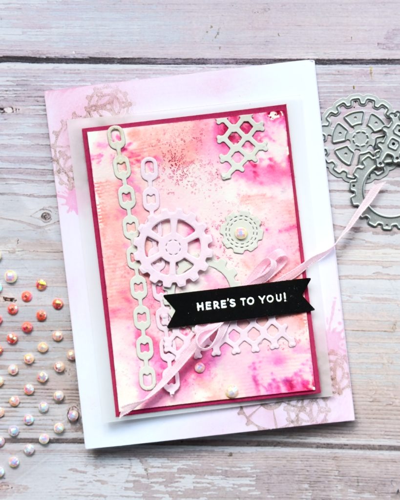A greeting card that has a pink water coloured background with different depths of pink There are chains, links and cogs layered on the top. The sentiment reads Heres to you