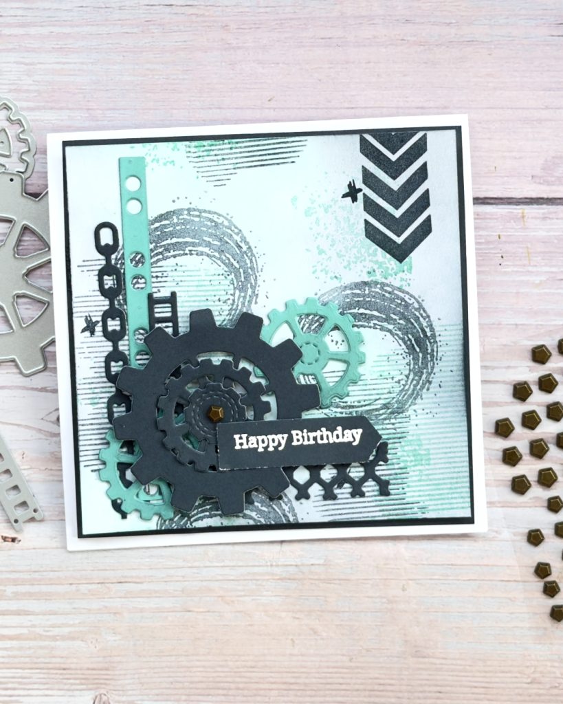 A square masculine Birthday Card with colours of black and teal, there are cogs stripes circles chains all layered on the card.
