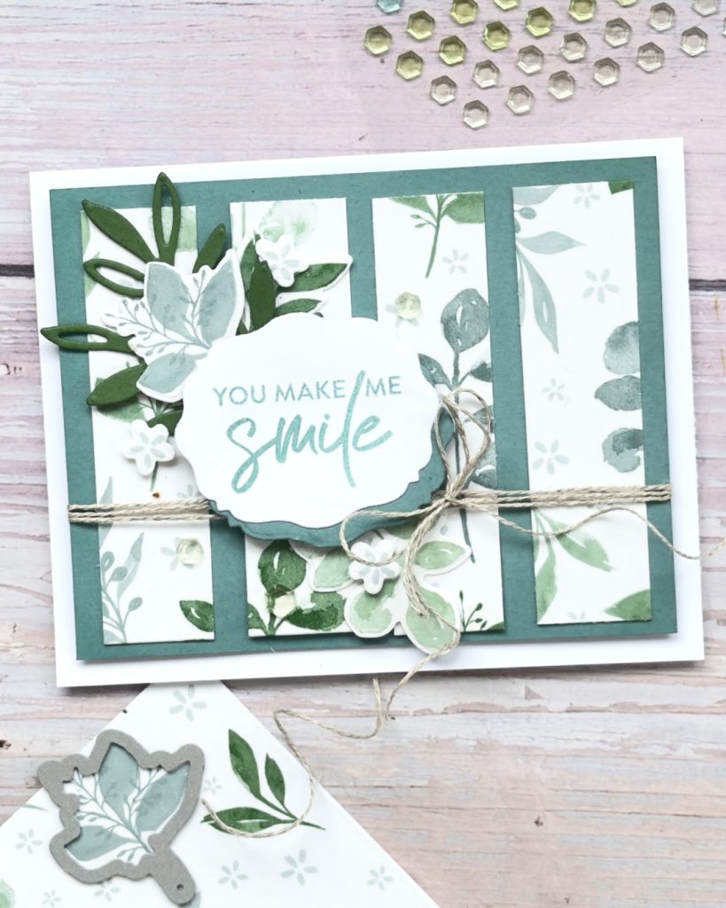 A handmade card featuring soft green botanical panels framed in dark green. Layered leaves and flowers surround a central label with the sentiment “You make me smile,” finished with twine tied across the card and small sequin accents.