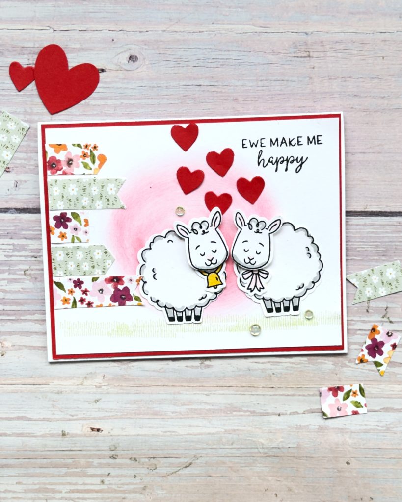 Handmade Valentine-style card using the Stampin’ Up Woolly Friends stamp and punch set, showing two stamped sheep, red felt hearts, soft pink ink blending, floral banner accents, and the sentiment “Ewe Make Me Happy.”