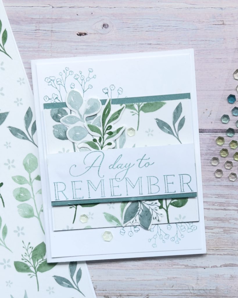 A handmade greeting card with soft green and sage botanical designs. Layered leaves and delicate floral images frame a central sentiment that reads “A day to remember.” The card is accented with subtle sequins and sits on a pale wooden background with coordinating patterned paper nearby.