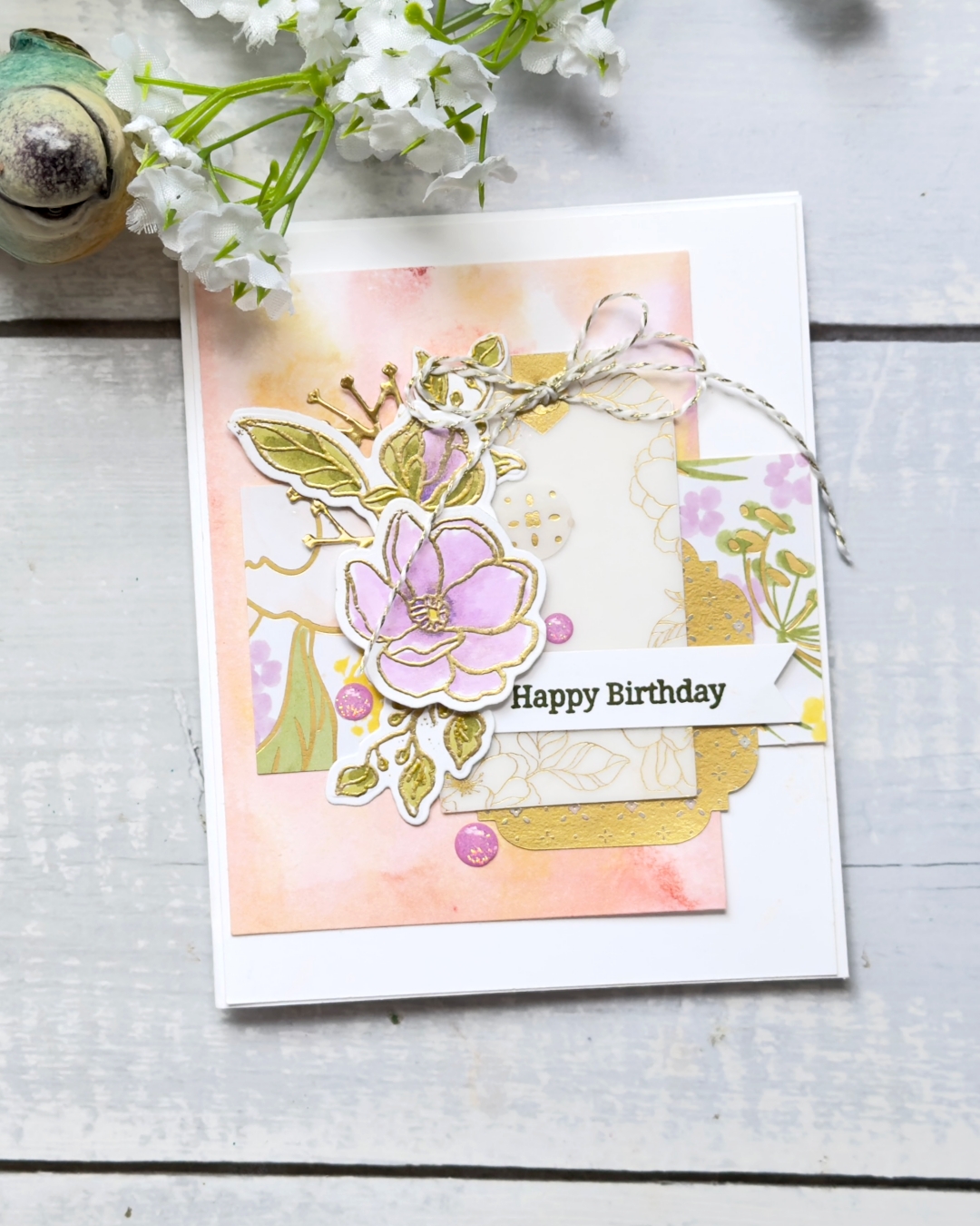 Handmade “Happy Birthday” card featuring a soft peach and pink alcohol-ink style background, layered with patterned paper, gold foil accents, and a gold-embossed purple floral focal image with green leaves. Finished with a white banner sentiment, pink embellishments, and a tied twine bow, photographed on a light wooden surface with white flowers above.
