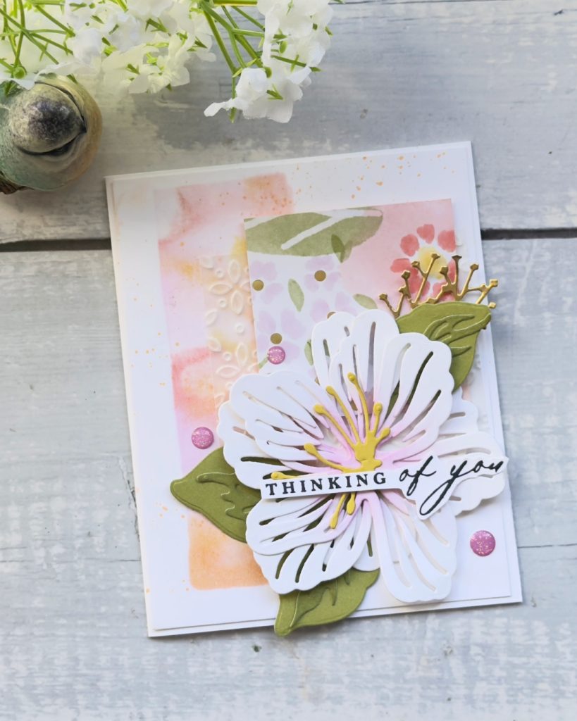 Handmade “Thinking of You” card featuring a layered white die-cut flower with soft pink and yellow details, olive green leaves, and gold accents. The background includes a pastel alcohol-ink style wash in peach and pink tones, embossed floral texture, and subtle splatter effects, photographed on a light wooden surface with white flowers above.